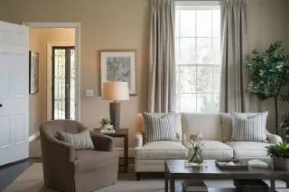 Un salon confortable avec un canapé beige, un fauteuil brun, une table basse, des coussins rayés et une grande fenêtre éclairant la pièce.