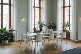 Une salle à manger lumineuse avec une grande table blanche entourée de chaises, baignée de lumière naturelle par de grandes fenêtres et agrémentée