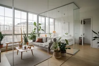 Un salon moderne et lumineux avec des plantes vertes et une cloison en verre séparant l'espace chambre.