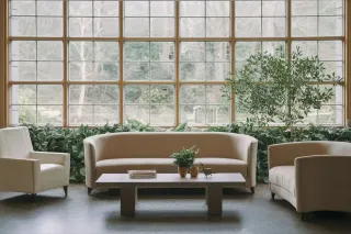 Un salon lumineux avec des fauteuils crème, une table basse orné de plantes vertes en pot, et une grande fenêtre offrant une vue sur le