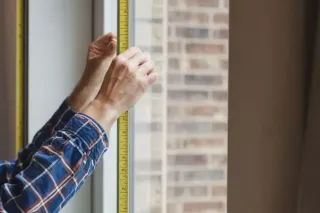 Une personne en chemise à carreaux mesure une fenêtre avec un ruban à mesurer.