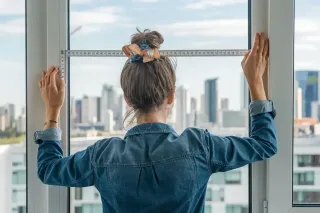 Une femme en chemise en jean mesure une fenêtre avec une règle, avec une vue sur les gratte-ciels en arrière-plan.