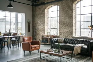 Un salon de style industriel avec des murs en briques, de grandes fenêtres et un mobilier moderne en cuir et en bois.