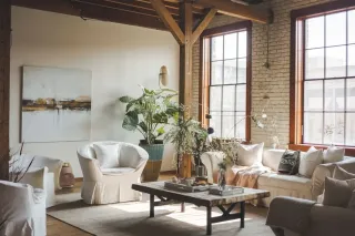 Un salon lumineux au style industriel avec mobilier blanc, grandes fenêtres, et plantes d'intérieur verdoyantes.