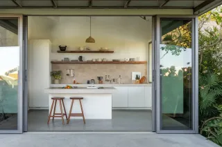 Cuisine moderne et minimaliste avec îlot central, tabourets en bois et grandes portes vitrées ouvertes sur l'extérieur.