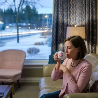 Une femme confortable assise dans un salon, dégustant une boisson chaude face à une fenêtre donnant sur un paysage hivernal.