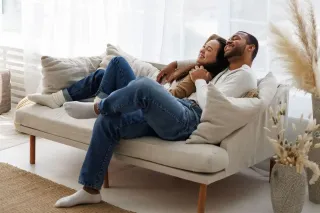 Un couple souriant est assis confortablement sur un canapé beige dans un salon lumineux et décoré de plantes.