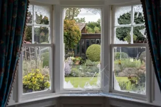 Vue d'un jardin serein à travers une fenêtre aux vitres brisées, encadrée de rideaux colorés.