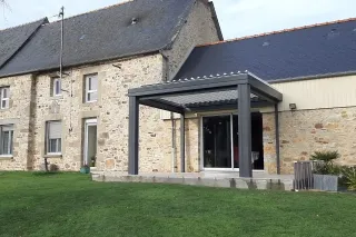 Une maison en pierre avec une pergola moderne en métal gris sur une pelouse verte.