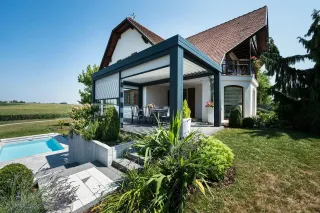 Maison moderne avec terrasse couverte et jardin verdoyant, intégrant une piscine et une vue sur la campagne environnante.