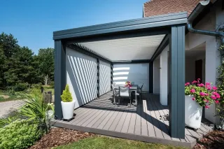 Une élégante pergola moderne en métal gris abrite une terrasse bois avec une table à manger entourée de plantes fleuries.