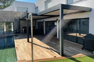 Terrasse moderne avec pergola en métal et piscine, donnant sur une maison contemporaine aux murs en pierre.