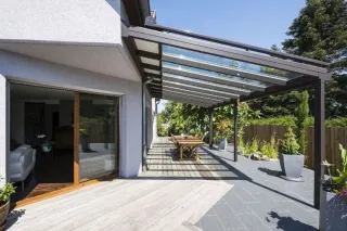 Terrasse moderne avec pergola en verre, mobilier de jardin en bois, et plantes luxuriantes, idéale pour profiter de l'extérieur.