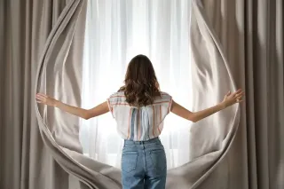 Une femme ouvre des rideaux beige pour laisser entrer la lumière du jour.