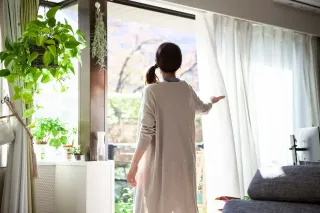 Une femme, de dos, ouvre les rideaux de sa maison entourée de plantes verdoyantes et baignées de lumière naturelle.