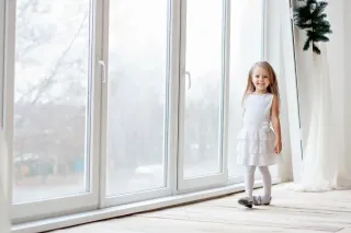 Une petite fille souriante en robe blanche se tient près d'une grande baie vitrée.