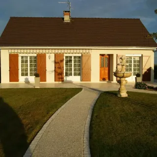 Une charmante maison de plain-pied avec volets en bois, allée en gravier et fontaine dans le jardin.