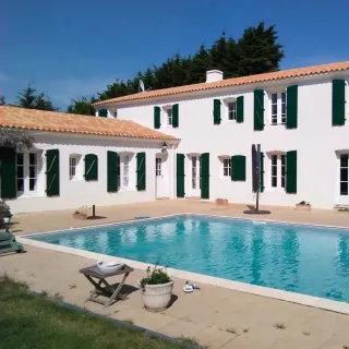 Belle maison blanche aux volets verts entourant une piscine dans un cadre ensoleillé.