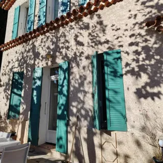 Façade ensoleillée d'une maison avec des volets bleus et des ombres d'arbres projetées sur le mur.