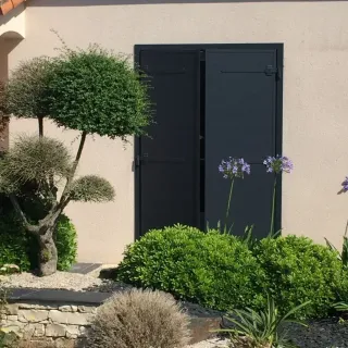 Façade de maison avec volets noirs fermés, entourée de plantes et d'un jardin bien entretenu.