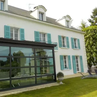 Grande maison blanche avec volets bleus et véranda moderne donnant sur un jardin verdoyant.