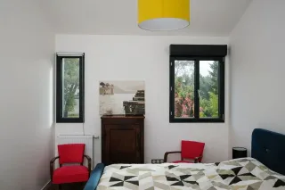 Une chambre lumineuse avec deux fenêtres, des fauteuils rouges, un lit avec une couverture géométrique, et une décoration murale apaisante