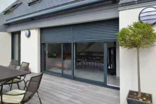 Façade de maison moderne avec grandes baies vitrées et volets roulants, donnant sur une terrasse en bois équipée de mobilier extérieur.