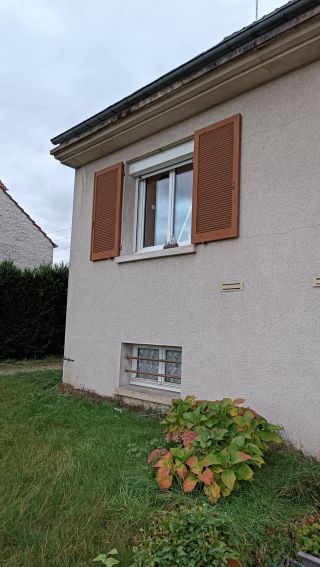 La photo montre un mur extérieur d'une maison avec une fenêtre munie de volets en bois et un petit jardin fleuri en premier plan.