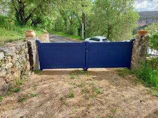 Une porte en bois bleue, entourée de murs en pierres avec des pots de fleurs, donne sur une allée verdoyante sous un ciel