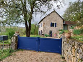 Maison en pierre traditionnelle avec volets bleus, entourée de verdure, derrière un portail bleu.