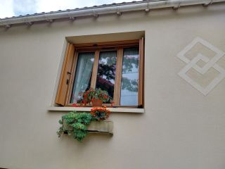 Une fenêtre en bois avec un pot de fleurs rouges et oranges sur le rebord, sur une façade beige avec un motif géométrique.