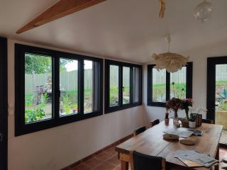 Une salle à manger lumineuse avec une grande table en bois, entourée de fenêtres donnant sur un jardin verdoyant.