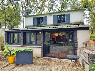 Petite maison avec de grandes baies vitrées entourée de verdure et de plantes en pot.