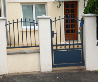 Portail en fer forgé bleu, encadré de piliers blancs, devant une entrée de maison avec une porte en bois vitrée.