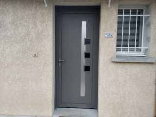 Une porte moderne en acier gris anthracite avec des éléments géométriques en verre, encadrée dans un mur en crépi beige.