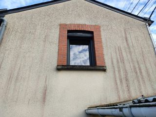 Façade d'une maison avec une fenêtre à encadrement en briques et des traces d'humidité sur le crépi beige.