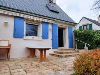 Une charmante terrasse en pierre donnant sur une maison avec des volets bleus et un toit en ardoise, entourée de végétation.