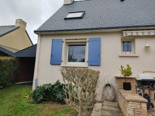 Une maison avec des volets bleus et un toit en ardoise, entourée d'un jardin bien entretenu avec un barbecue et des plantes