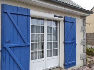 Une porte-fenêtre blanche avec des volets bleus sur un mur en crépi, entourée de pierres et d'un petit jardin.