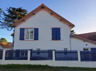 Maison traditionnelle avec volets bleus et toit en tuiles, entourée d'une clôture en bois, sous un ciel dégagé.