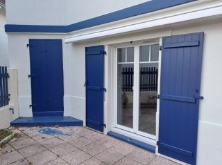 Façade de maison avec des volets bleus et une porte-fenêtre donnant sur une terrasse en pierre.