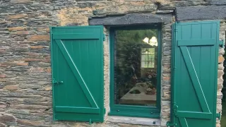 Une fenêtre à volets verts ouverts, encadrée de pierre rustique, offre une vue sur l'intérieur d'une maison chaleureuse.