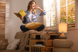 Une femme assise près d'une fenêtre ensoleillée lit un livre tout en tenant un coussin jaune sur ses genoux.