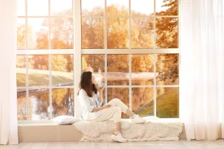 Une femme assise sur un banc devant une grande fenêtre, entourée d'un paysage d'automne, savourant une tasse de café.