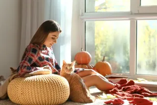 Une jeune femme assise sur un tapis, caressant un chat, avec des citrouilles en arrière-plan près d'une fenêtre éclairée par la