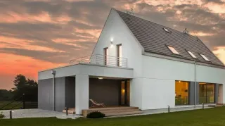 Maison moderne blanche avec un toit gris, éclairée en soirée par un ciel orangé.