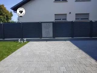 Clôture moderne en métal gris foncé avec un panneau décoratif central et une allée pavée devant une maison.