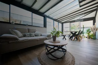 Une véranda lumineuse avec de grandes baies vitrées, un canapé beige, une table basse ronde et une table à manger en bois, décor