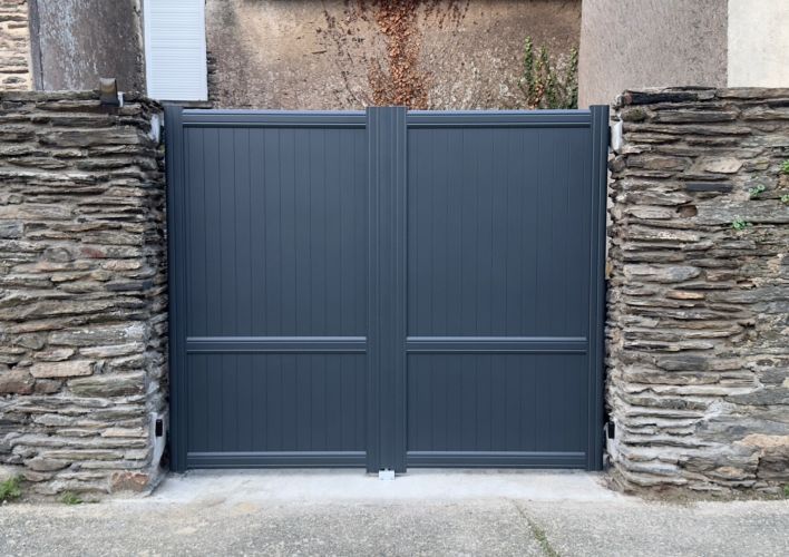 Un portail moderne en métal gris anthracite s'ouvre entre deux piliers en pierre rustique.