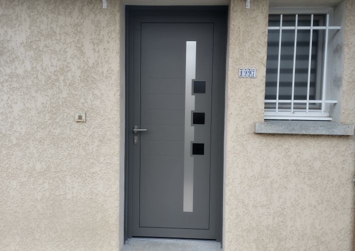 Une porte moderne en acier gris anthracite avec des éléments géométriques en verre, encadrée dans un mur en crépi beige.